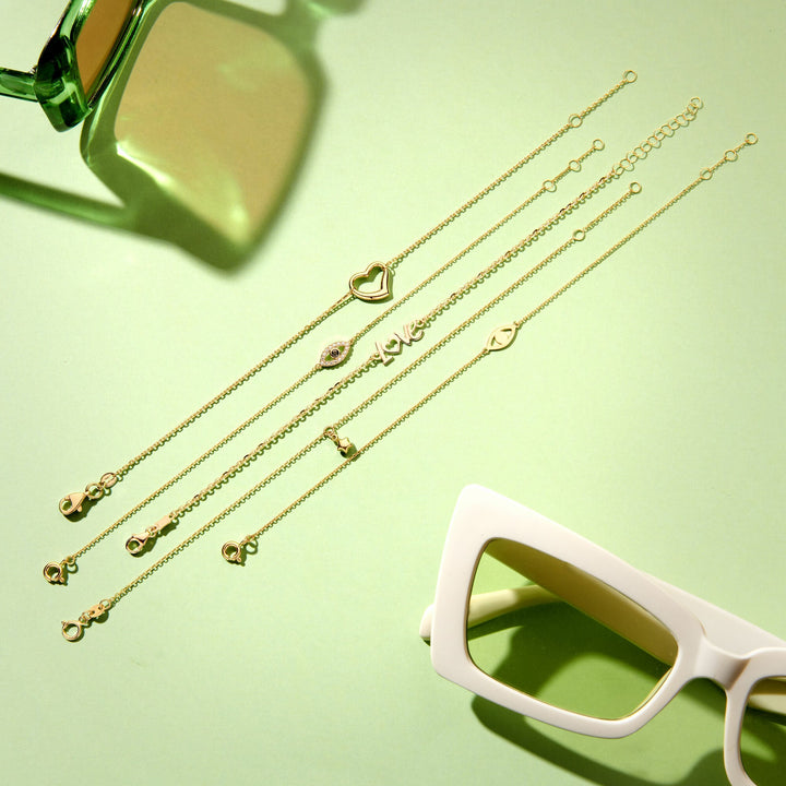 Vijf Van der Kooij Collectie Gouden armband dames hart 14K gouden armbanden met verschillende bedels zijn diagonaal gerangschikt op een pastelgroen oppervlak, geaccentueerd door groene en witte zonnebrillen in de hoeken.