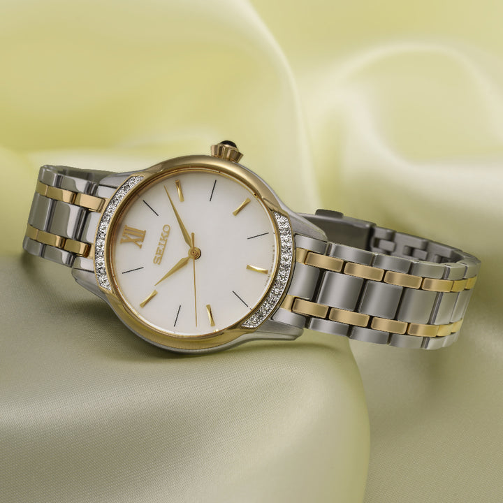 Het Seiko dameshorloge SRZ544P1 heeft een zilveren en gouden metalen band, een witte wijzerplaat met gouden markeringen en wijzers, omlijst door een roestvrijstalen kast. Fonkelende stenen sieren de lunette, allemaal afgezet tegen een zachtgele stoffen achtergrond voor een elegante touch.