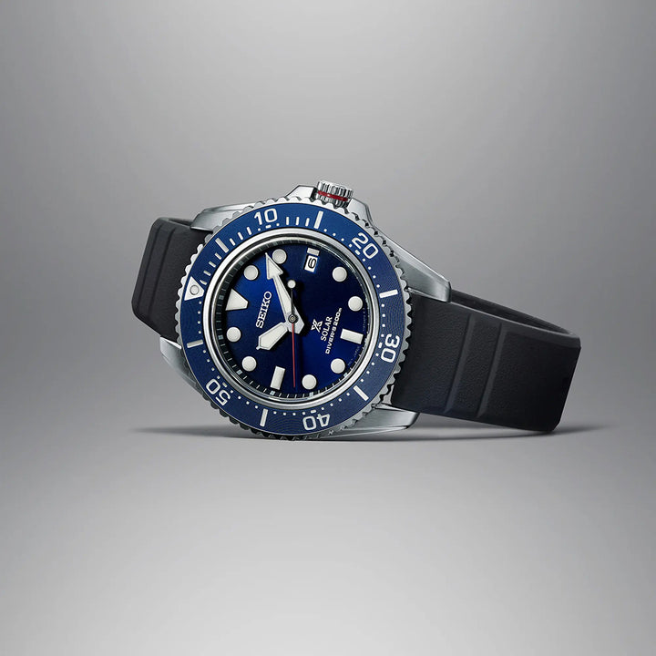 Het Seiko Prospex heren horloge SNE593P1 heeft een blauwe wijzerplaat, opvallende witte markeringen, draaibare lunette, datumfunctie op 3 uur en wordt gecombineerd met een zwarte rubberen band, afgezet tegen een grijze achtergrond.