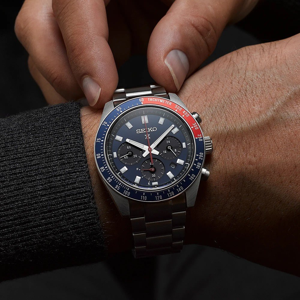 Een persoon draagt een Seiko Prospex SSC913P1 chronograafhorloge van roestvrij staal, met een zwarte wijzerplaat, blauwrode lunette, subdials en tachymetermarkeringen. Ze passen het op zonne-energie werkende uurwerk op hun pols aan.