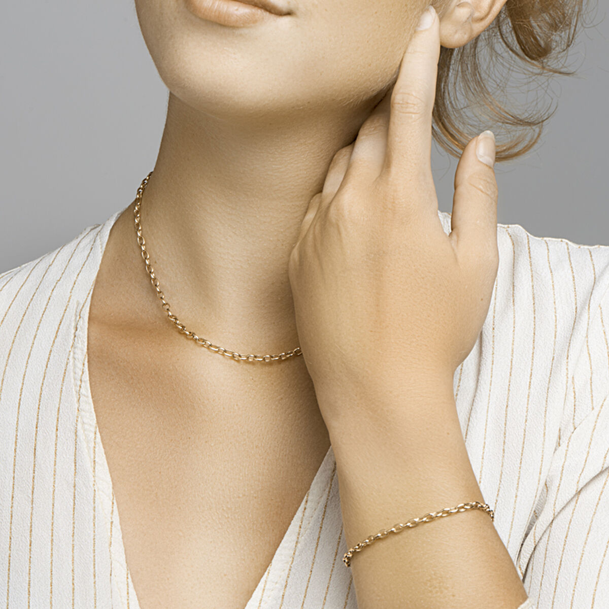 Een vrouw in een witte krijtstreepblouse showt een delicaat gouden kettinkje en de Gouden armband dames anker 14K van Van der Kooij Collectie, haar hand zachtjes in haar nek.