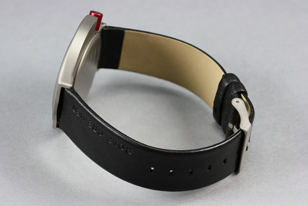Het Rolf Cremer Horloge 492314, een unisex kwartshorloge van Rolf Cremer, heeft een zwarte leren band met een zilverkleurige gesp, een tan bandinterieur en een rode knop op de kast, weergegeven op een lichtgrijs oppervlak.