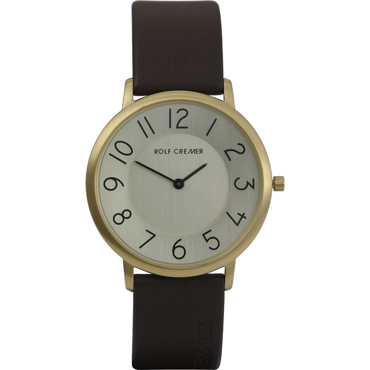 Het Rolf Cremer Horloge 503706 is een minimalistisch quartzhorloge met een bruine leren band, ronde gouden kast en zilveren wijzerplaat. Het heeft zwarte Arabische cijfers in een modern lettertype, slanke zwarte wijzers en het merk Rolf Cremer elegant gedrukt in het midden, wat een tijdloze stijl weerspiegelt.