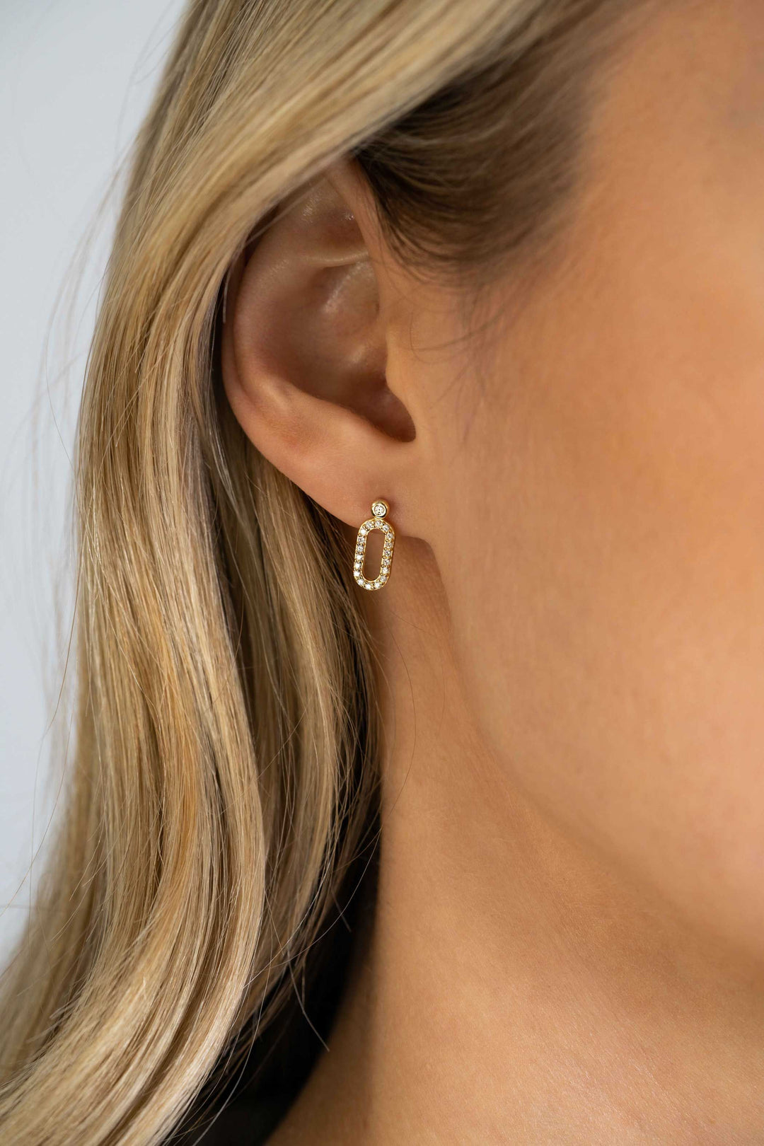 Close-up van het oor van een vrouw met Zinzi Oorbellen Gold Plated Zio2398 Y hoepel oorbel, met een ovale vorm met ingebedde zirkonia stenen en een enkele ronde steen aan de bovenkant, perfect passend bij haar lichtblonde haar.
