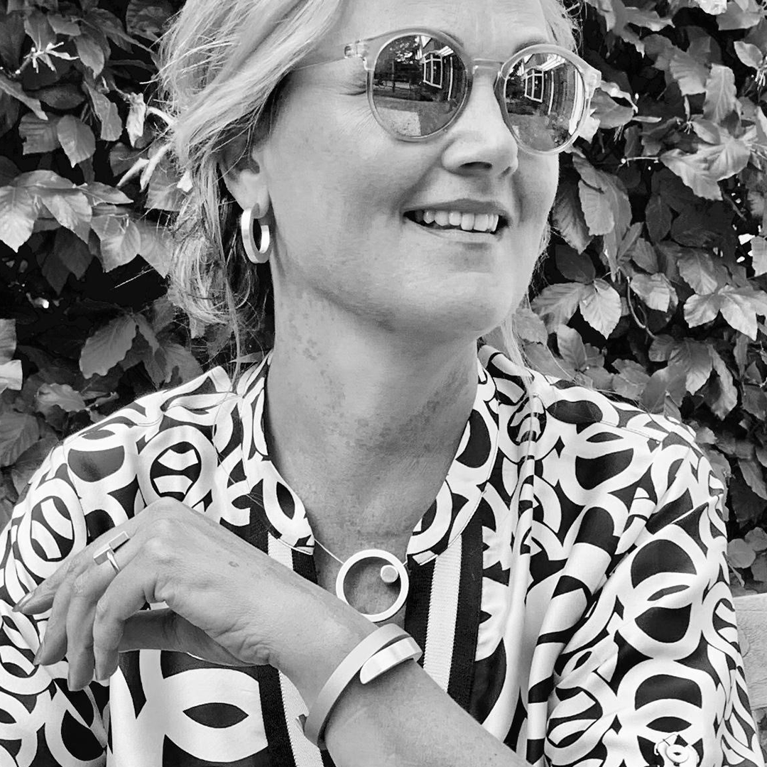 Een vrouw in een blouse met patroon en een zonnebril glimlacht terwijl ze voor een bladerrijke achtergrond zit. Ze draagt ronde oorbellen, een ring met een magnetische sluiting en een Armband Cs gehecht Rood A1R armband van CLIC Creations. De zwart-witfoto straalt een stijlvolle, relaxte sfeer uit.