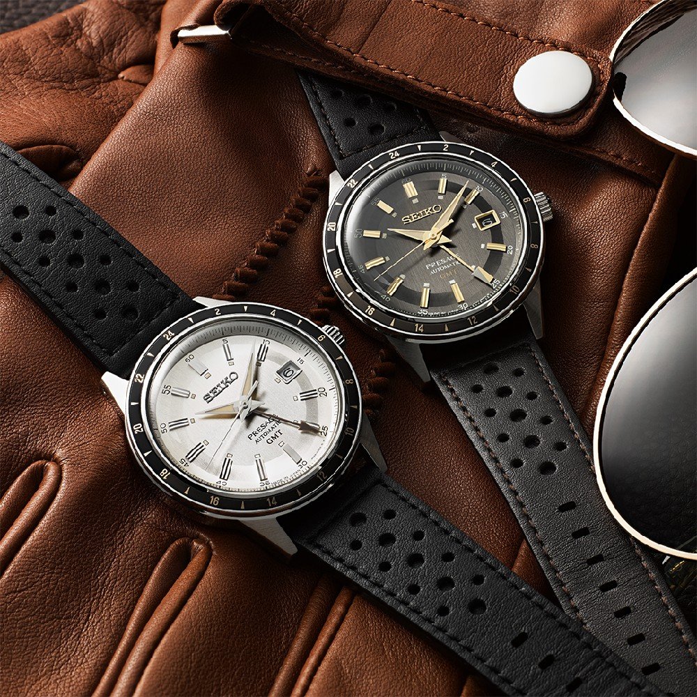 Twee Seiko Presage heren horloges, met zwarte leren bandjes, rusten op een bruine leren handschoen. De ene heeft een witte wijzerplaat, de andere een zwarte. Zonnebrillen met metalen montuur voegen subtiele elegantie toe.