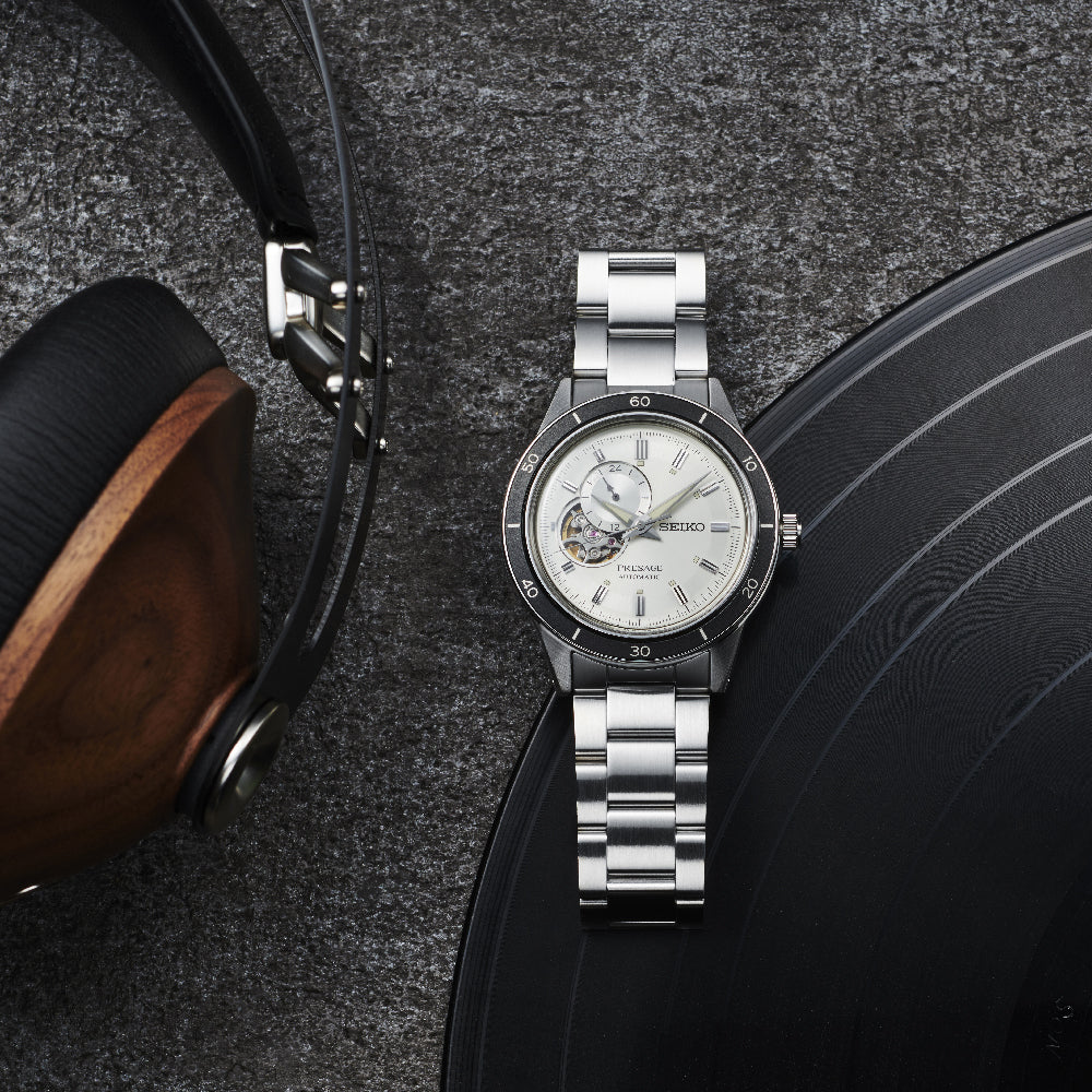 De Seiko Presage heren horloge SSA423J1, met een witte wijzerplaat en open hart ontwerp, rust op een donker oppervlak naast grote houten-koptelefoon met een deel van een zwarte vinylplaat in de buurt. Het horloge heeft een roestvrijstalen kast en metalen armband.