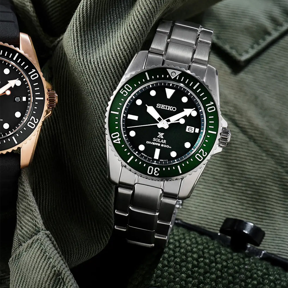 Een close-up van de Seiko Prospex heren horloge SNE583P1, een duikhorloge op zonne-energie, heeft een groene lunette en een roestvrijstalen band op een gestructureerde stof. De wijzerplaat toont de datum met gedetailleerde minutenmarkeringen en biedt 20ATM waterdichtheid, met een ander horloge gedeeltelijk zichtbaar.