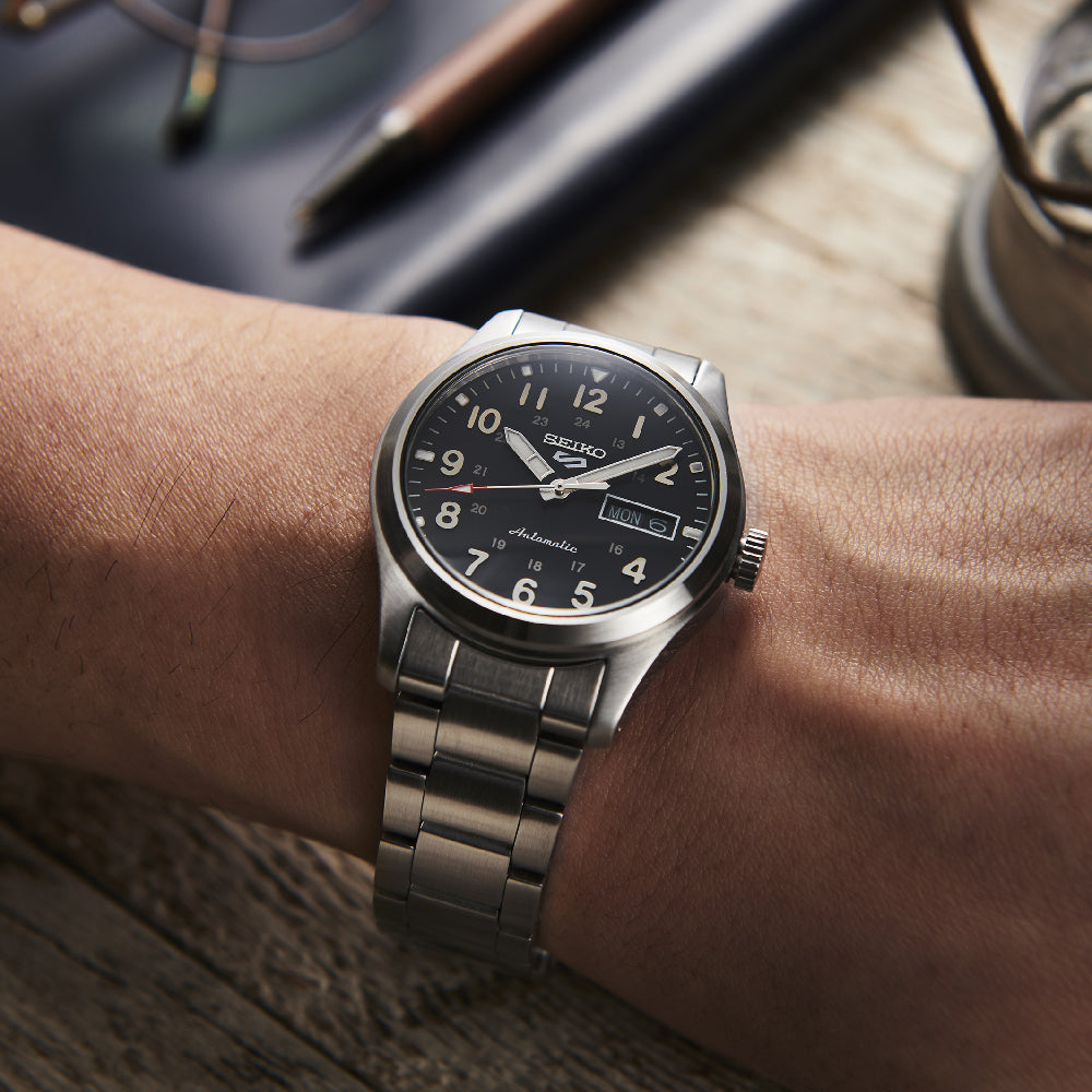 Close-up van een pols met een Seiko 5 Sports heren horloge SRPG27K1, met een automatisch roestvrijstalen ontwerp met een zwarte wijzerplaat en zilveren uurmarkeringen. Een datumweergave is zichtbaar. De achtergrond toont een houten oppervlak met een pen en papier.