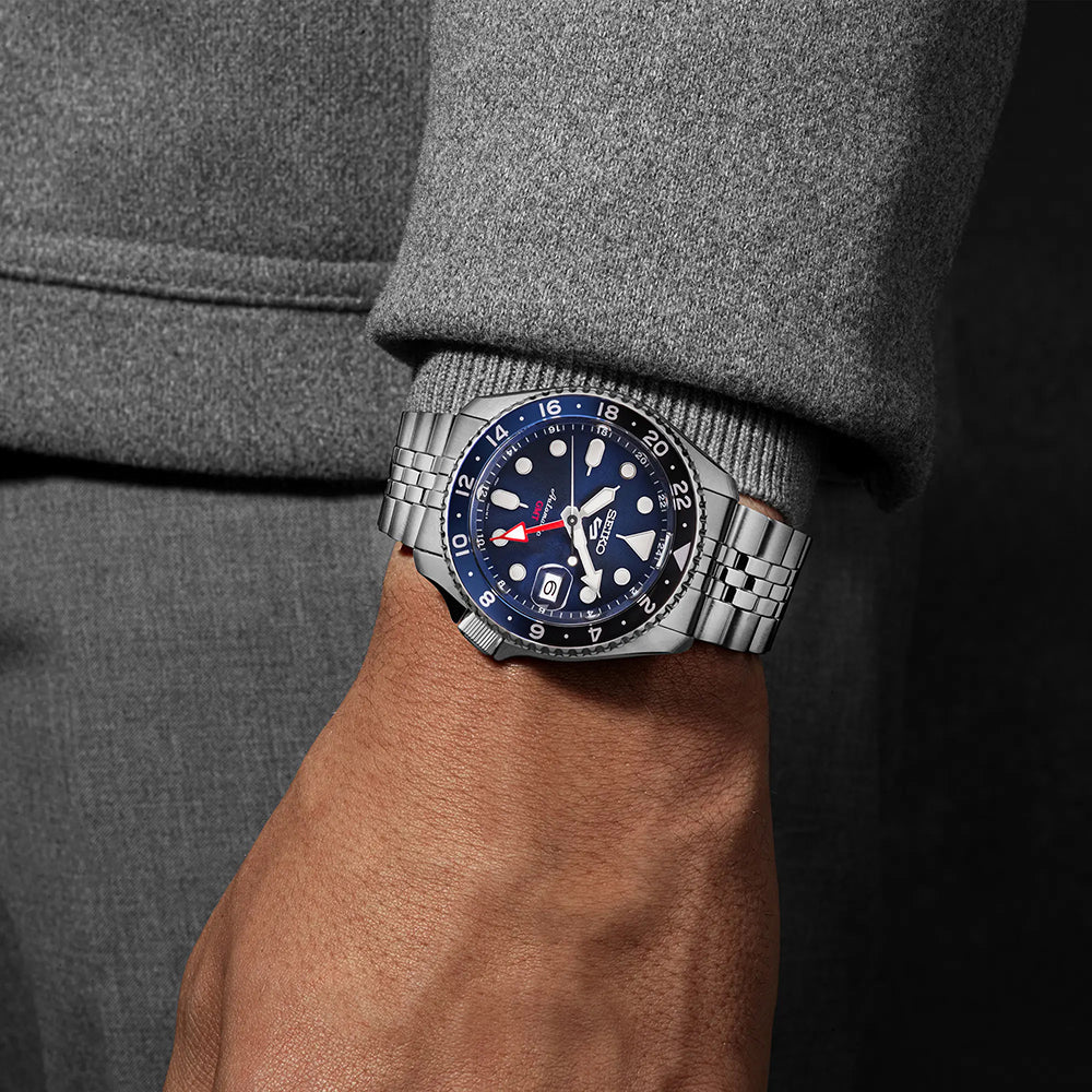 Een persoon draagt het Seiko 5 Sports heren horloge SSK003K1 van Seiko, een automatisch horloge met een blauwe wijzerplaat en rood/witte accenten. Dit zilveren metalen horloge is waterbestendig tot 10ATM en vormt een elegante aanvulling op een grijs shirt met lange mouwen, terwijl hun hand nonchalant naast hen rust.