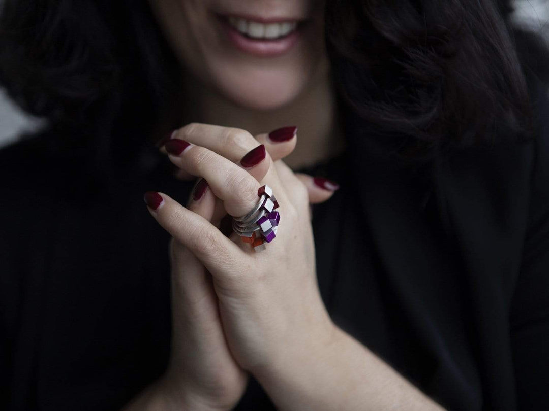 Een persoon met donker haar en bordeauxrode nagels klapt in zijn handen, toont een kleurrijke Ring Kubusjes Zwart R4Z van CLIC Creations - een verstelbaar, handgemaakt sieraad uit Nederland - en glimlacht zachtjes.