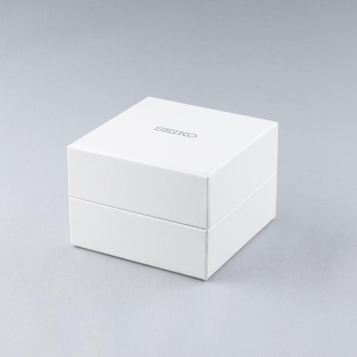 Een witte, vierkante Seiko-kast met een matte afwerking en SEIKO in metalen letters op de bovenkant, geeft blijk van de precisie van het Seiko herenhorloge SUR421P1 met quartz uurwerk aan de binnenkant, afgezet tegen een effen grijze achtergrond.