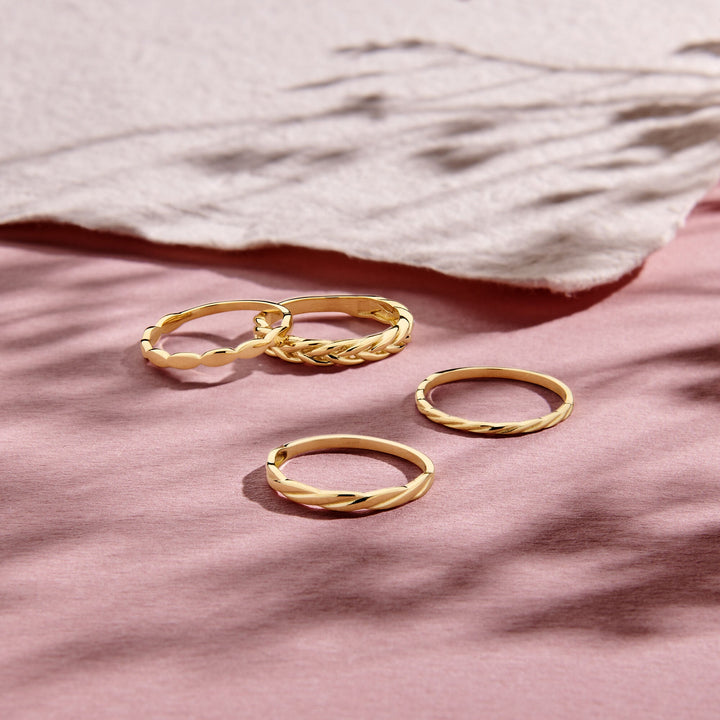 Vier gouden ringen, waaronder de Van der Kooij Collectie ring gedraaid 14K geelgoud en een klassieke 14k geelgouden ring, worden tentoongesteld op een zachtroze oppervlak met gestructureerd crèmepapier en zachte schaduwen erboven.