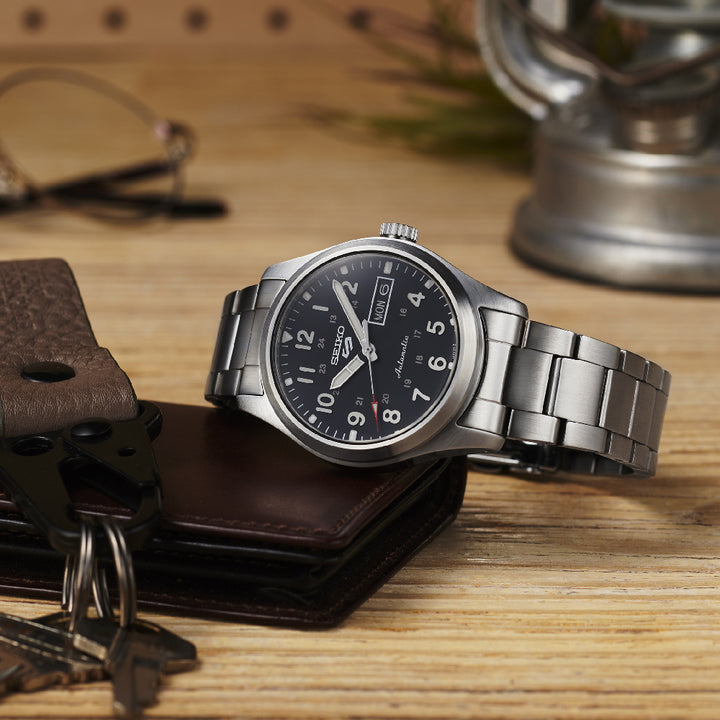 Een Seiko 5 Sports herenhorloge SRPG27K1 met een zilveren metalen band en zwarte wijzerplaat rust op hout, naast een leren portemonnee en een set sleutels, onder het zachte licht van een lamp. Perfect stijlvol.