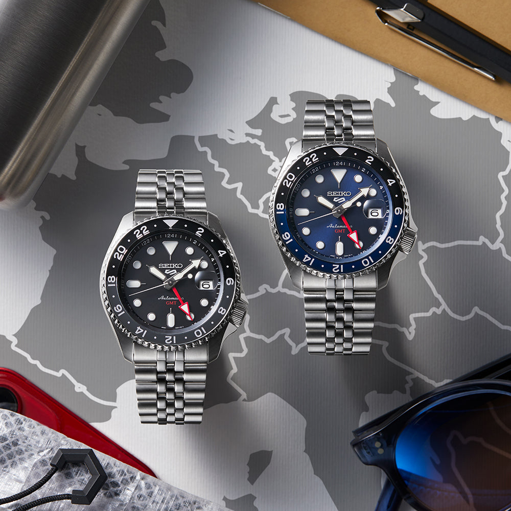 Twee Seiko 5 Sports herenhorloges met zilverkleurige metalen banden rusten op een grijs-witte wereldkaart. De ene heeft een zwarte wijzerplaat met een rode secondewijzer; de andere is het Seiko 5 Sports herenhorloge SSK003K1 met een blauwe wijzerplaat en blauw-zwarte lunette.