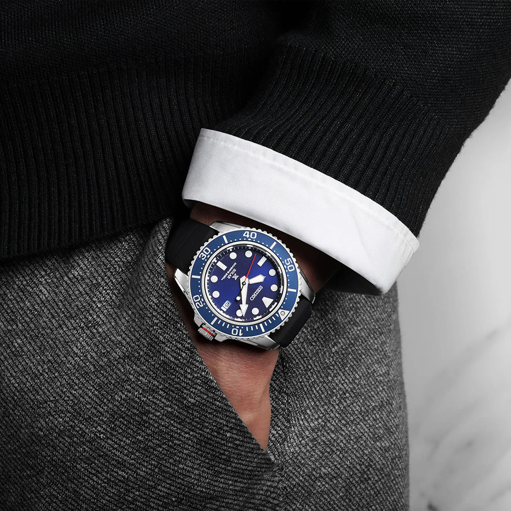 Een persoon draagt casual een stijlvol Seiko Prospex heren horloge SNE593P1 met een opvallende blauwe wijzerplaat en prominente markeringen. Het duikhorloge, met een zwarte band, complementeert hun donkere trui en witte overhemd terwijl hun hand moeiteloos in hun zak rust.