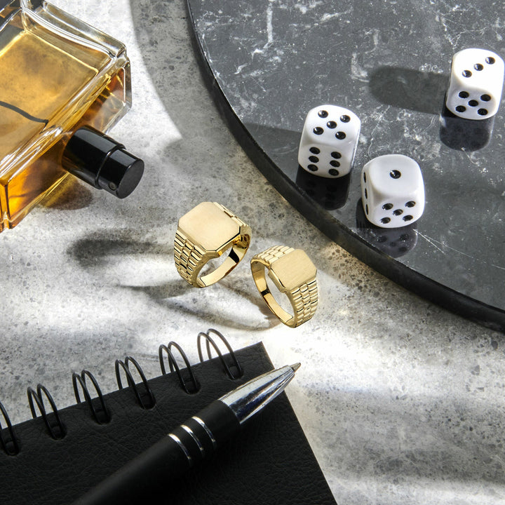 Twee Van der Kooij Collectie graveerringen gematteerd 14K geelgoud, een zwarte pen op een spiraalvormig notitieboekje, drie witte dobbelstenen op een rond zwart marmeren dienblad en een amberkleurig parfumflesje liggen op een gestructureerd grijs oppervlak in natuurlijk licht.