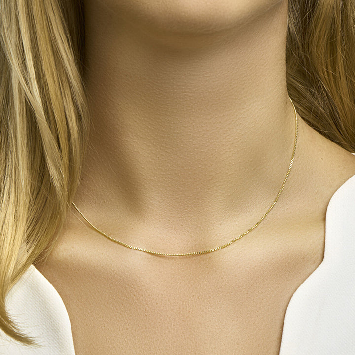 Een vrouw met lichtblond haar en een witte top draagt de Van der Kooij Collectie collier gourmette 4-zijdes geslepen 1,2 mm 14K geelgoud. De afbeelding benadrukt haar nek en borst, waardoor het subtiele gastronomische ontwerp goed naar voren komt.