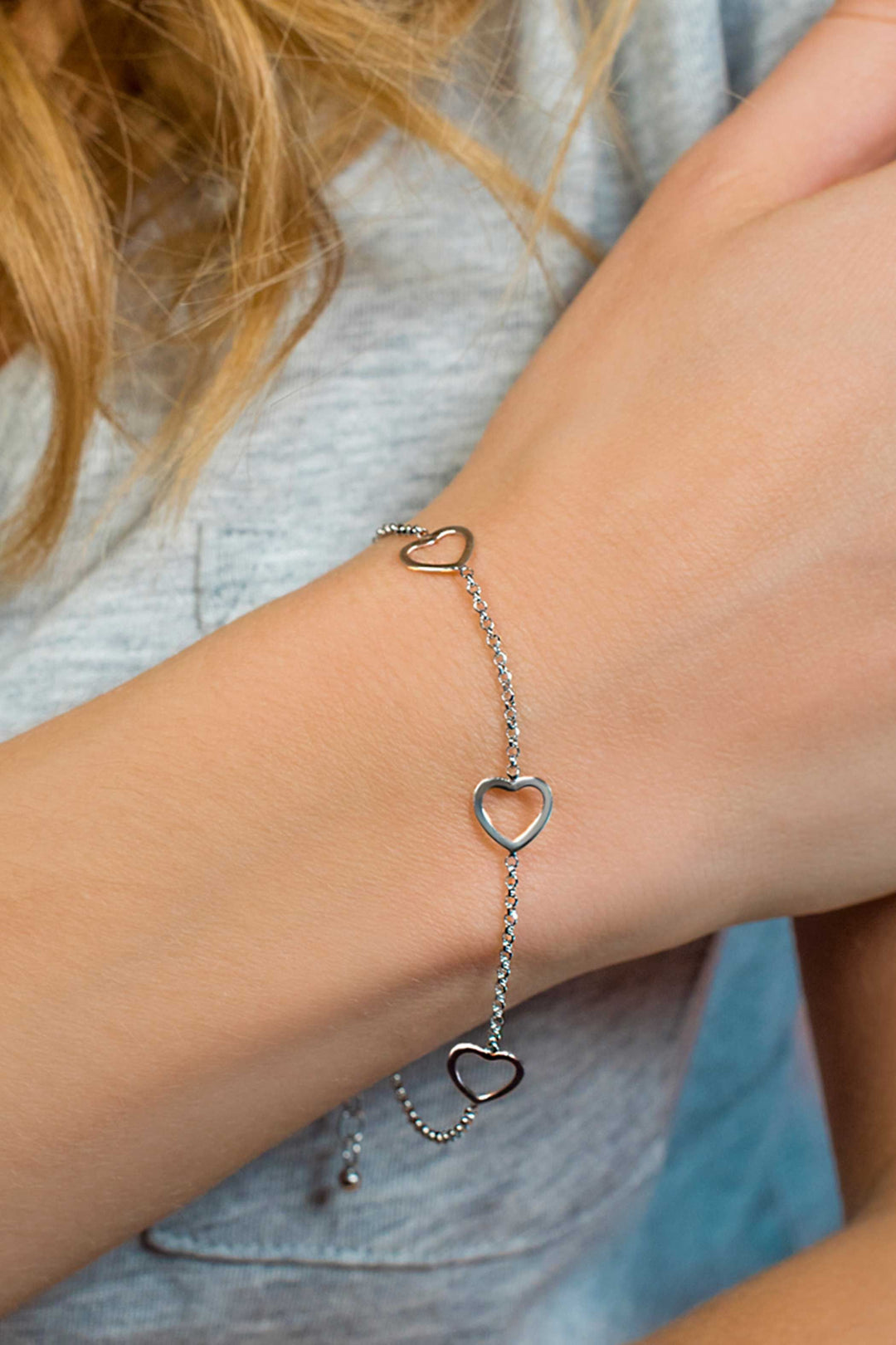 De Zinzi Armband Zilver Zia1188 met delicate hartvormige schakels siert de pols van een persoon. Het elegante stuk, met een afmeting van 17-20 cm, verbetert hun lichtgrijze bovenkant, terwijl lichtbruin haar charme toevoegt aan het ensemble.