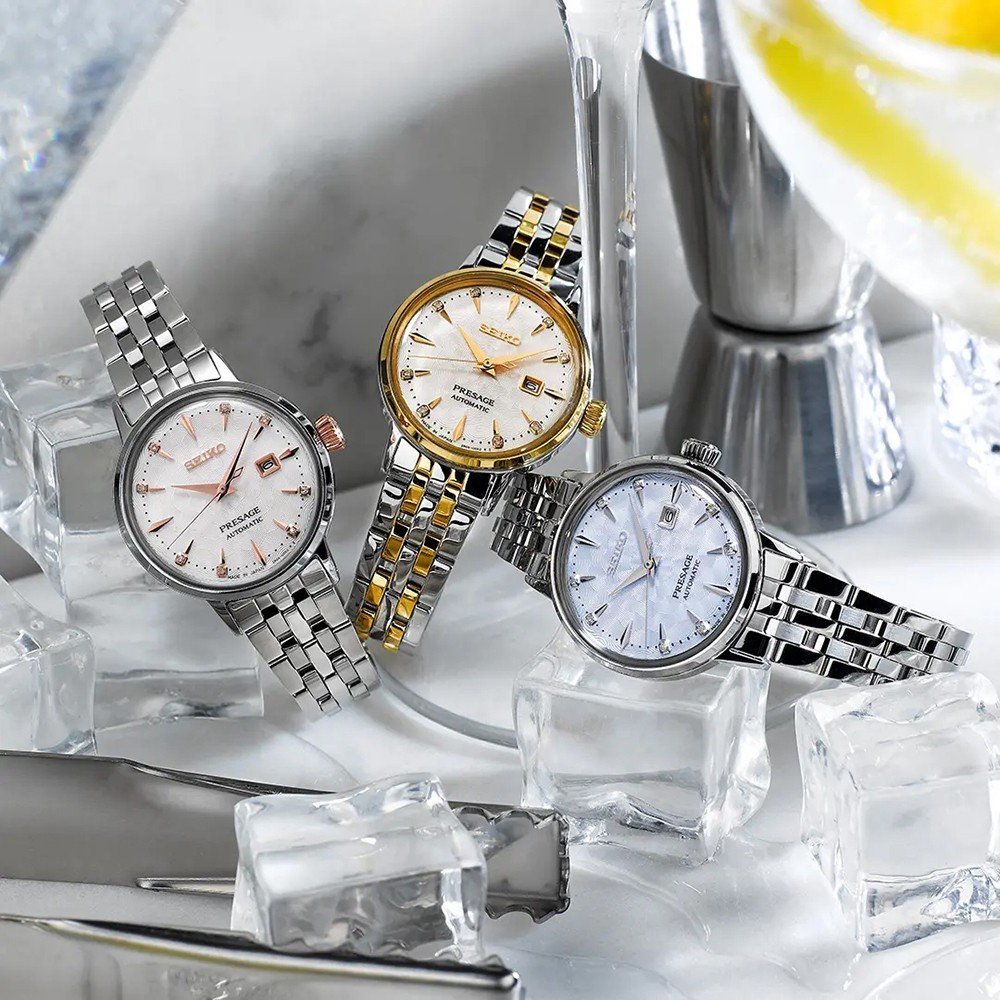 Drie Seiko polshorloges met metalen banden, waaronder de Seiko Presage dames horloge SRE010J1, staan rechtop tussen ijsblokjes en cocktailgereedschap. Een zilver-en-goudkleurig model springt eruit, met een schijfje citroen op de achtergrond.