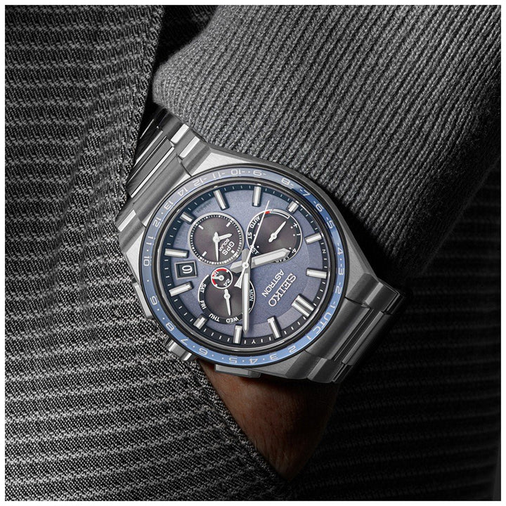 De Seiko Astron SSH109J1 herenhorloge heeft een donkergrijze wijzerplaat met meerdere wijzerplaten, waaronder een datumweergave en chronograaf, beschermd door robuust saffierglas. Gestyled met een getextureerd grijs pak, rust de hand van de drager in de zak.
