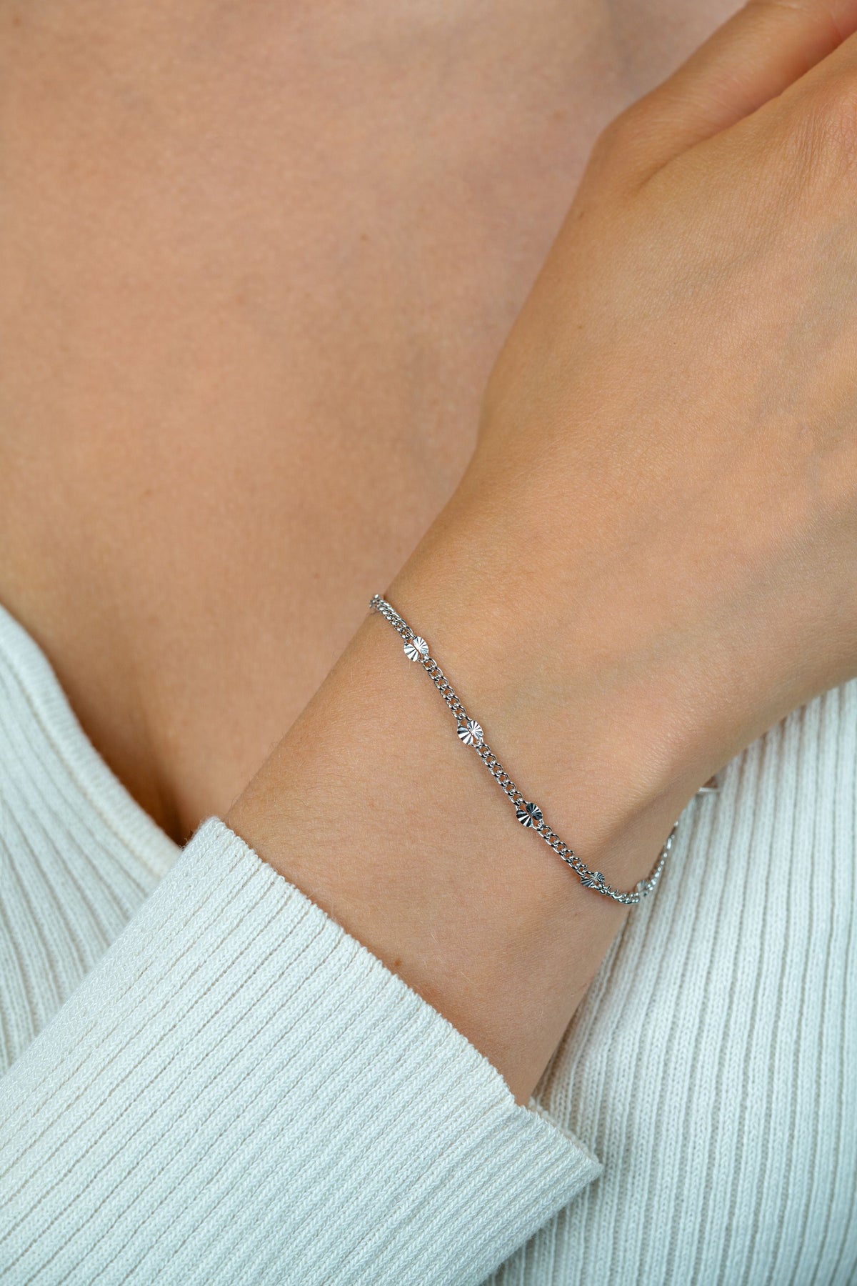 Een close-up van een pols die de Zinzi Armband zilver ZIA2713 van Zinzi draagt, een delicaat eerste gehalte zilveren gourmet schakelarmband met kleine bedeltjes en rodiumlaag, afgebeeld tegen een lichtbeige geribbelde top bij het sleutelbeen.