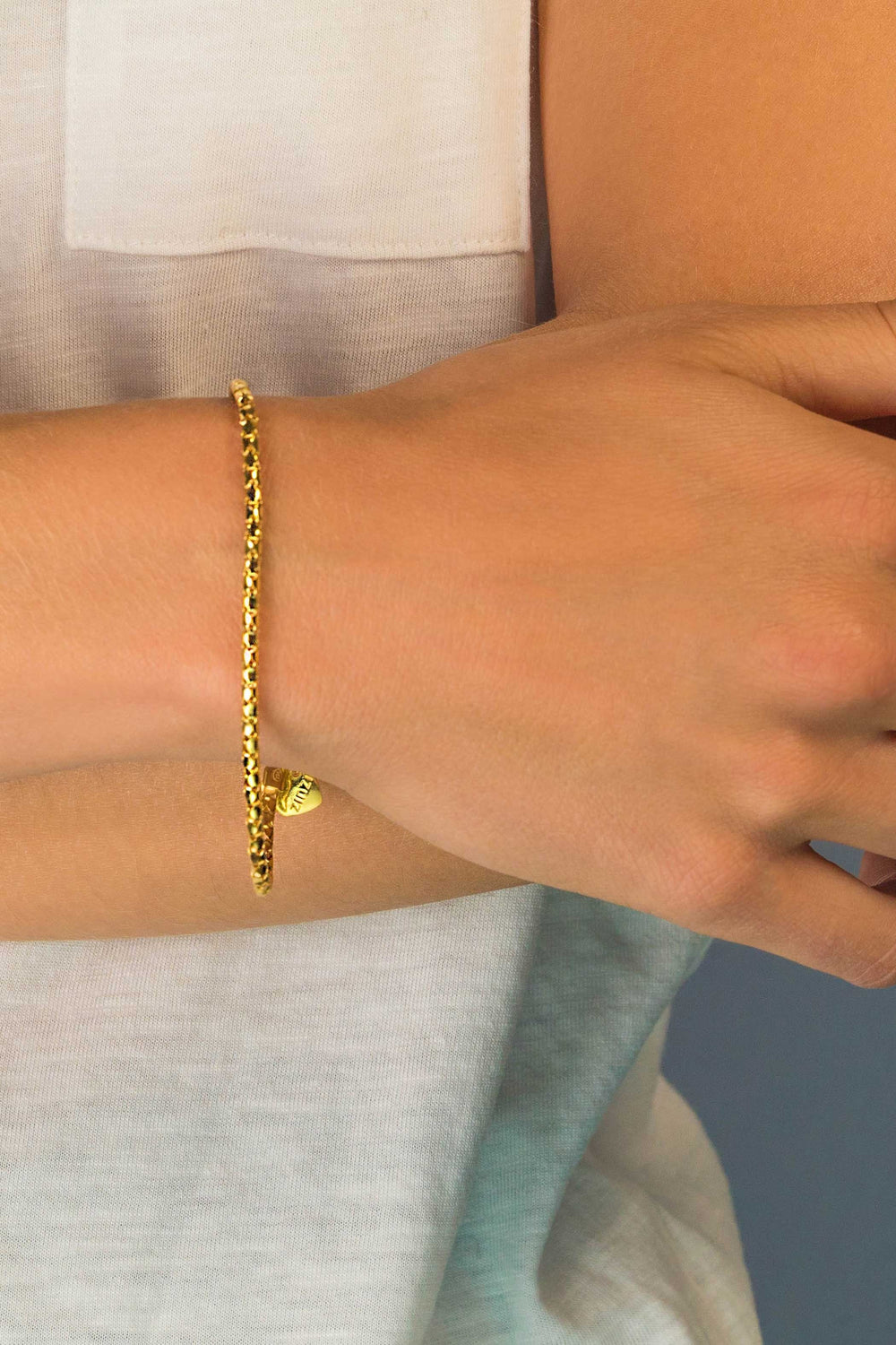 Een close-up van de arm van een persoon toont de Zinzi Armband Gold Plated Zia1129 Ga, met een delicate gouden ketting en een klein hartje. Ze dragen een wit mouwloos topje en houden hun arm in een ontspannen houding tegen een neutrale achtergrond, wat moeiteloze elegantie uitstraalt.