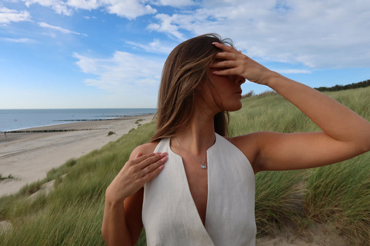 Een vrouw in een mouwloze witte jurk staat in het hoge gras bij het strand, draagt het Amélie AsHanger massief van Juwelier van der Kooij, met één hand op haar borst en de andere haar gezicht beschermend onder een gedeeltelijk bewolkte hemel.
