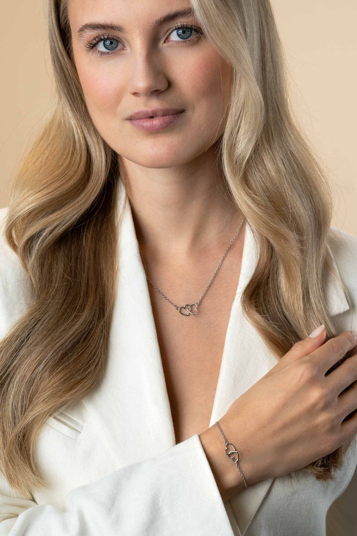 Een vrouw met lang blond haar en een vriendelijke glimlach draagt een witte blazer en de Zinzi Ketting Zilver Zic2513 van Zinzi, gecombineerd met een bijpassende zilveren hart armband, poserend tegen een neutrale achtergrond.