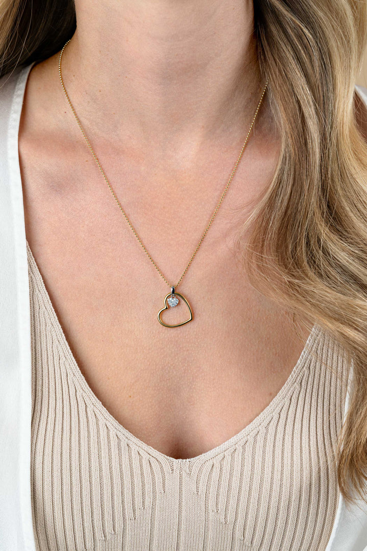 Een vrouw in een beige geribbelde top en een wit shirt draagt de Zinzi ketting ZIC2621 45-48cm, een gouden hartvormige hanger met een klein edelsteenaccent, van Zinzi, van dichtbij rond haar nek en sleutelbeen.