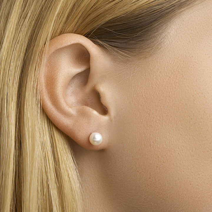 Een close-up toont een vrouw met steil blond haar en Van der Kooij Collectie oorknoppen parel 14K geelgoud – eenvoudige ronde oorknopjes met gladde zoetwaterparels op haar oorlel. De afbeelding is helder belicht, waardoor de parel en het goud goed uitkomen.