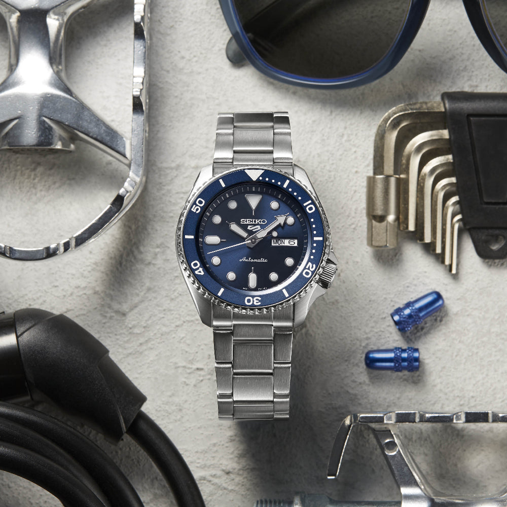 De Seiko 5 Sports heren horloge SRPD51K1, een zilveren horloge met een blauwe wijzerplaat en metalen band, wordt weergegeven op een gestructureerd oppervlak, vergezeld door een zonnebril, een moersleutel, inbussleutels, een kabel en twee blauwe doppen. Dit automatische horloge toont elegant zowel de tijd als de datum.