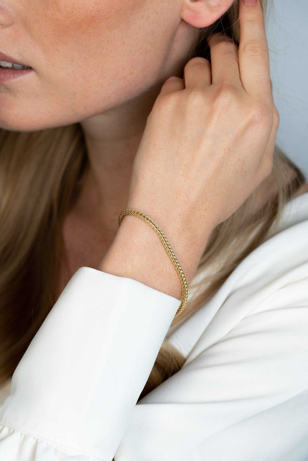 Een close-up toont iemand met lang haar in een witte blouse die zijn haar aanraakt. De pols is voorzien van een delicate Zinzi Armband Gold Plated Zia1925 G, die de zachte, neutrale achtergrond van de scène versterkt.