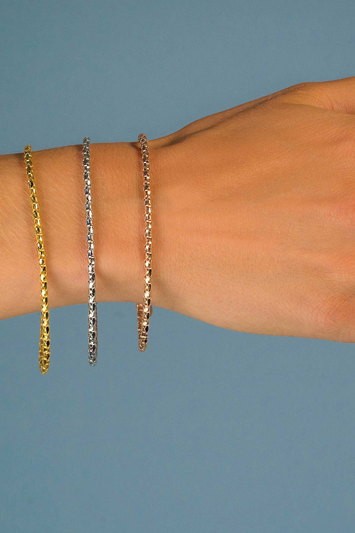 Een hand versierd met drie tennisarmbanden in geel, wit en roségoud siert een effen blauwe achtergrond. De betoverende gloed van de Zinzi Armband Gold Plated Zia1129 Ga voegt een vleugje elegantie toe aan deze verbluffende weergave.