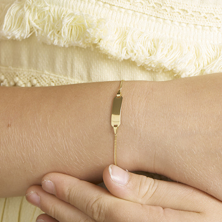 Een close-up van een pols versierd met de Van der Kooij Collectie graveerarmband gourmet plaat 5 mm (11-13 cm), vingers zachtjes rustend op de pols, tegen crèmekleurige structuurstof met franje details.
