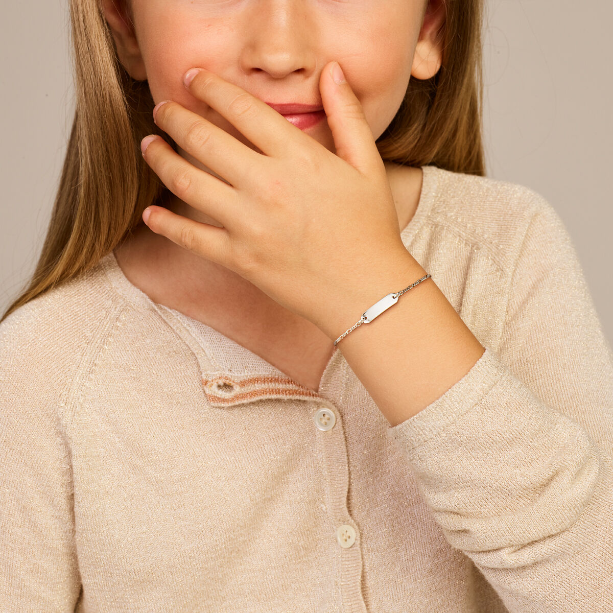 Een jong meisje met lang lichtbruin haar glimlacht zachtjes, haar hand bedekt gedeeltelijk haar mond. Ze draagt een beige trui en de Van der Kooij Collectie graveerarmband plaat 5,0 mm 11 + 2 cm zilver gerhodineerd om haar pols voor een subtiele elegante touch.