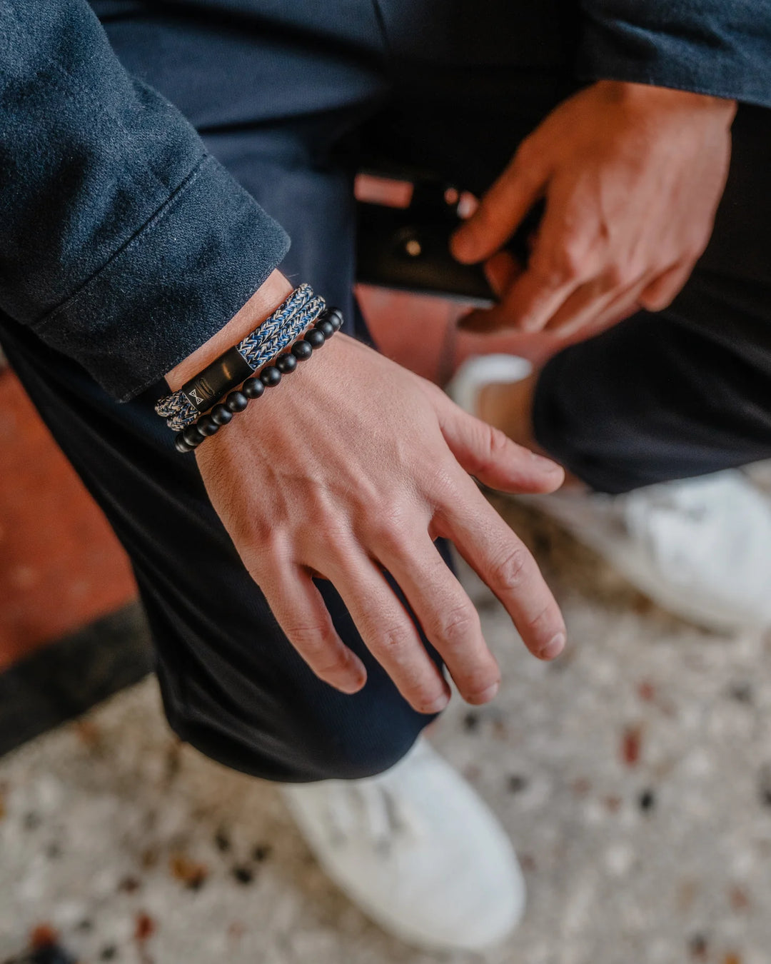 Zittend toont een persoon in een marineblauwe jas en zwarte broek zijn linkerhand versierd met armbanden met Heren Edelstenen kralen. Witte sneakers raken een gevlekte vloer, wat doet denken aan het ruige terrein van Black Elk Peak. De Kralen armband van AZE Jewels is gemaakt met 8mm stenen.