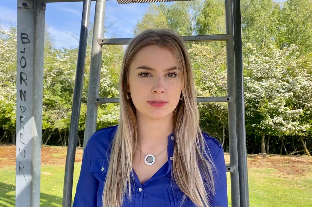 Een vrouw met lang blond haar, in een blauw shirt en een Collier Rondje in een ring C12 ketting van CLIC Creations, staat buiten. Achter haar zijn groene bomen en een metalen constructie met tekst. De lucht is helder en blauw.