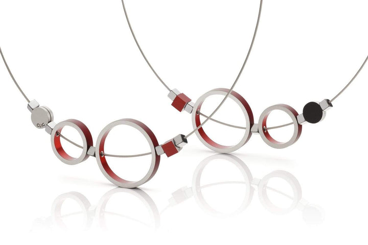 De Collier Ringen Rood C186R van CLIC Creations heeft een minimalistisch ontwerp met onderling verbonden cirkels en vierkanten. Metalen elementen hebben rode accenten en zijn verbonden op dunne draadkettingen met een magnetische sluiting, allemaal tegen een overzichtelijke witte achtergrond voor een moderne artistieke flair.
