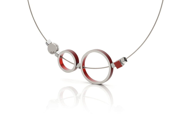 De Collier Ringen Rood C186R van CLIC Creations is een moderne ketting met een uniek ontwerp van twee metalen cirkels met rode accenten die lijken op een bril, hangend aan een dunne draadketting. Een subtiel logo siert de kleine ronde bevestiging aan de linkerkant, die allemaal prachtig reflecteren op een wit oppervlak.