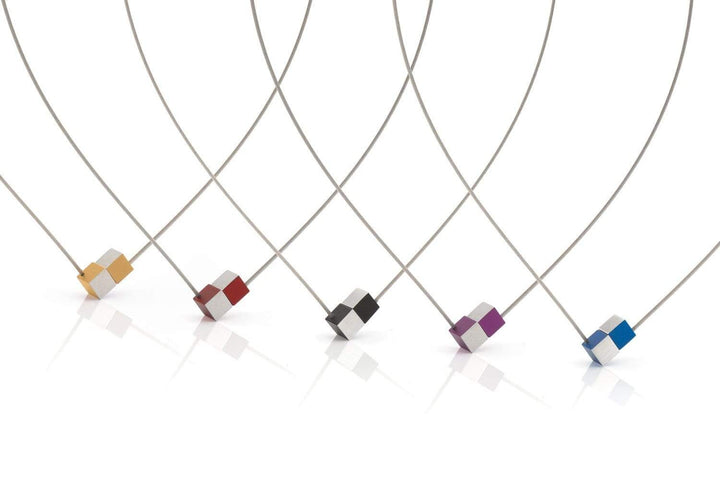 Vijf moderne CLIC Creations-kettingen hebben dunne draden en kleine geometrische kubushangers in unieke kleuren geel, oranje, rood, zwart, paars en blauw. Handgemaakt in Nederland met een chique design en geplaatst tegen een schone witte achtergrond.