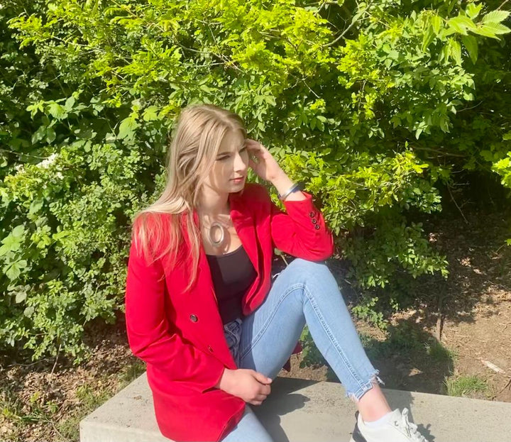 Een vrouw met lang blond haar zit op een betonnen richel bij weelderige groene struiken, gekleed in een rode blazer, blauwe jeans en witte sneakers. Ze draagt de Collier Grote ring met Ovaal C242 van CLIC Creations met een uniek ontwerp en magnetische sluiting. Haar blik dwaalt zijwaarts terwijl haar hand bij haar gezicht rust.