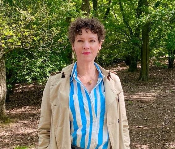 Een persoon met kort krullend haar staat buiten, stijlvol gekleed in een beige trenchcoat over een blauw-wit gestreept shirt, aangevuld met het unieke ontwerp van CLIC Creations Collier Ovaaltjes tegen elkaar Goud|Geel C245G. Dit handgemaakte in Nederland ensemble harmonieert met de rustige parkachtige achtergrond.