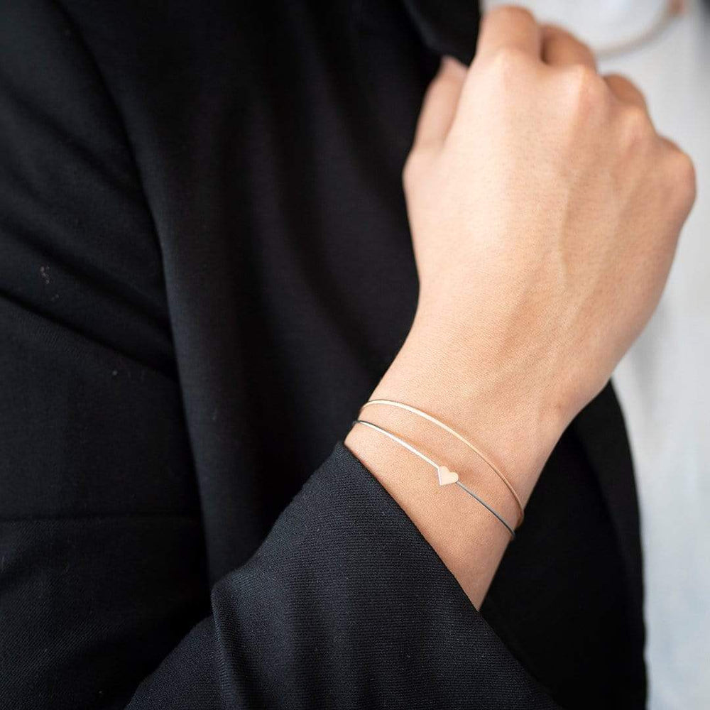 Een persoon in een zwart jasje, hand op kin, draagt de Thinking of You ❤️ armband goud van CLIC Creations. De delicate armband met een klein hartje is handgemaakt in Nederland, en valt op tegen de wazige achtergrond.