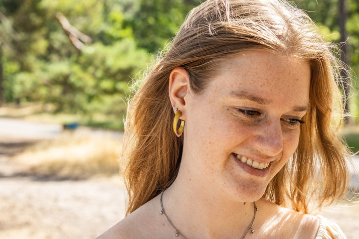 Een persoon met lang haar glimlacht terwijl hij naar beneden kijkt, staande buiten in een zonnige natuurlijke omgeving met bomen en gras. Ze dragen hypoallergene ovale creolen van CLIC Creations en een ketting van chirurgisch edelstaal.
