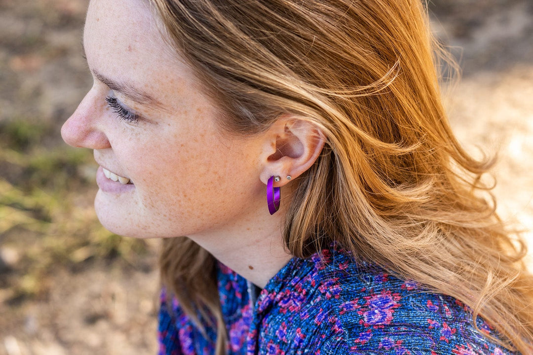 Een vrouw met lang haar lacht terwijl ze CLIC Creations Ovale creolen Paars O21P draagt, gemaakt van chirurgisch staal. Haar blauwe kledingstuk met roze patronen complementeert de kleine, paarse hoepeloorbellen. De achtergrond is een wazige natuurlijke setting.