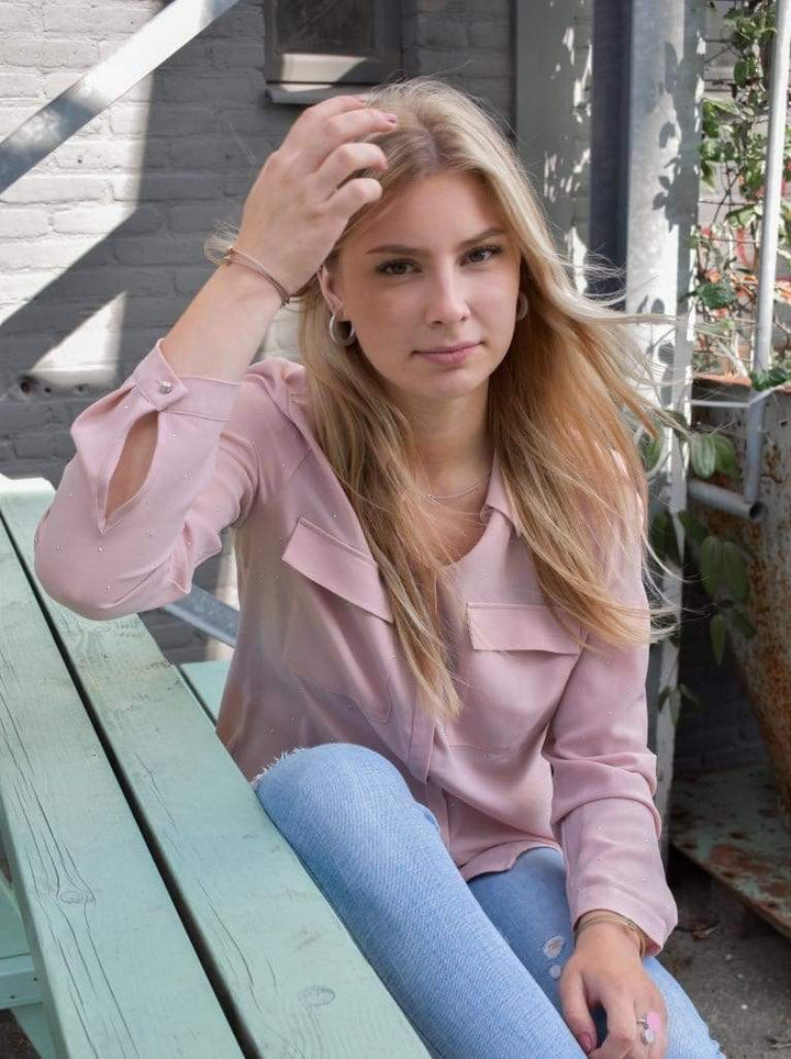 Een jonge vrouw met lang blond haar zit op een mintgroene bank, gekleed in een lichtroze blouse en jeans. Haar serene uitdrukking past bij het groen achter haar. Ze draagt CLIC Creations Creolen Ronde ringen Zwart O10Z oorbellen, wat een uniek tintje toevoegt aan haar chique look tegen de bakstenen muur als achtergrond.