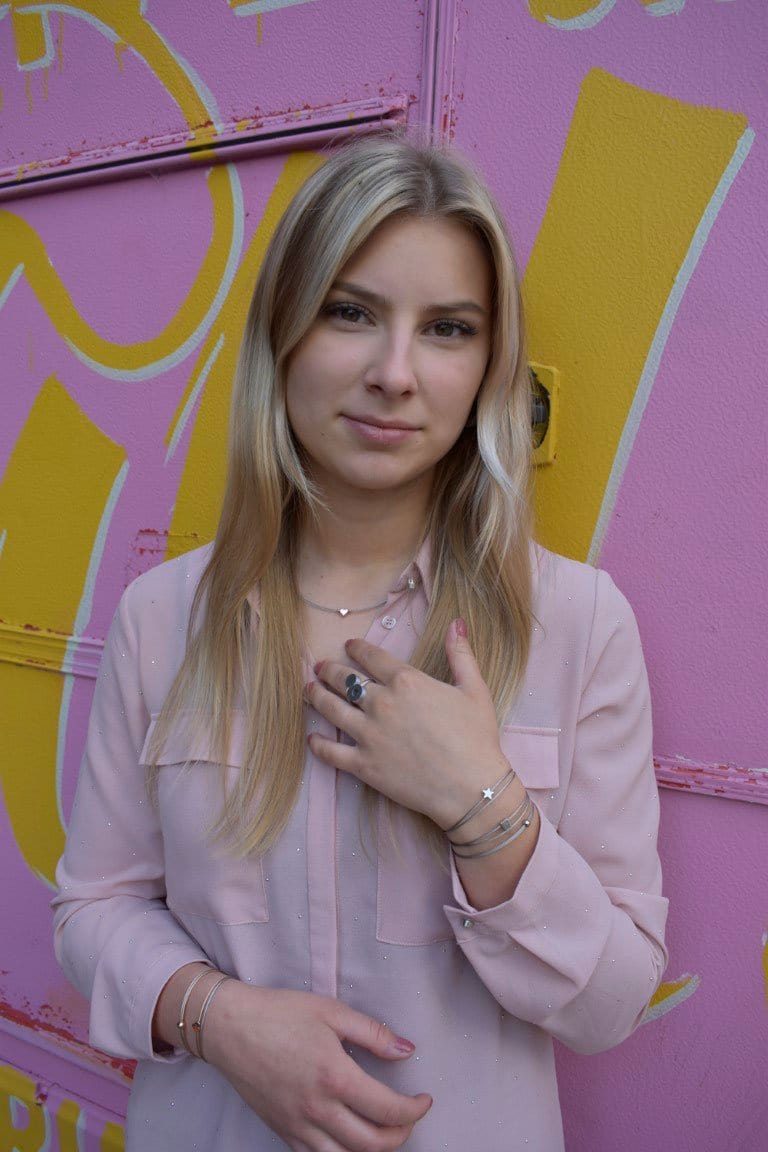 Een persoon met lang blond haar staat voor een roze en gele muur, draagt een lichtroze blouse en meerdere armbanden, waaronder een CLIC Creations Thinking of You 🦋 armband goud. Eén hand raakt hun borst aan terwijl ze in de camera kijken, en legt de essentie van een persoonlijk cadeau vast.