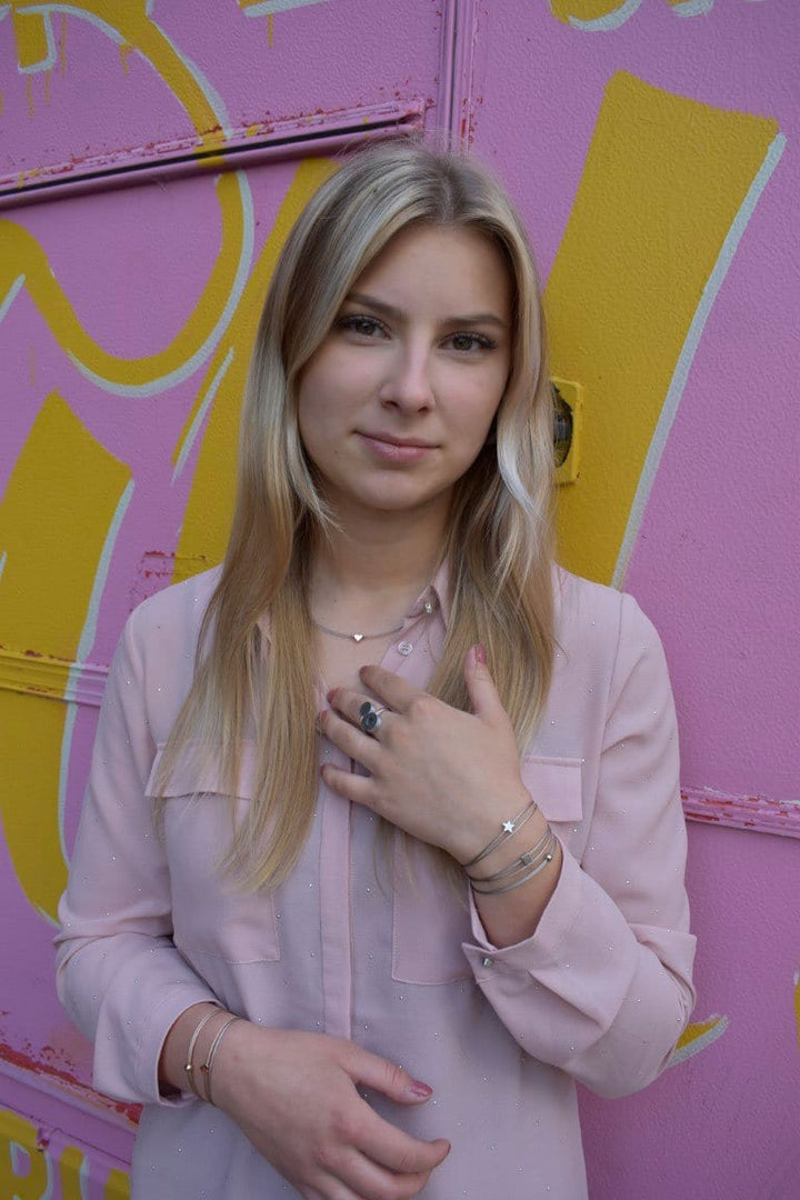 Een persoon met lang blond haar staat voor een roze en gele muur, draagt een lichtroze blouse en meerdere armbanden, waaronder een CLIC Creations Thinking of You 🦋 armband goud. Eén hand raakt hun borst aan terwijl ze in de camera kijken, en legt de essentie van een persoonlijk cadeau vast.