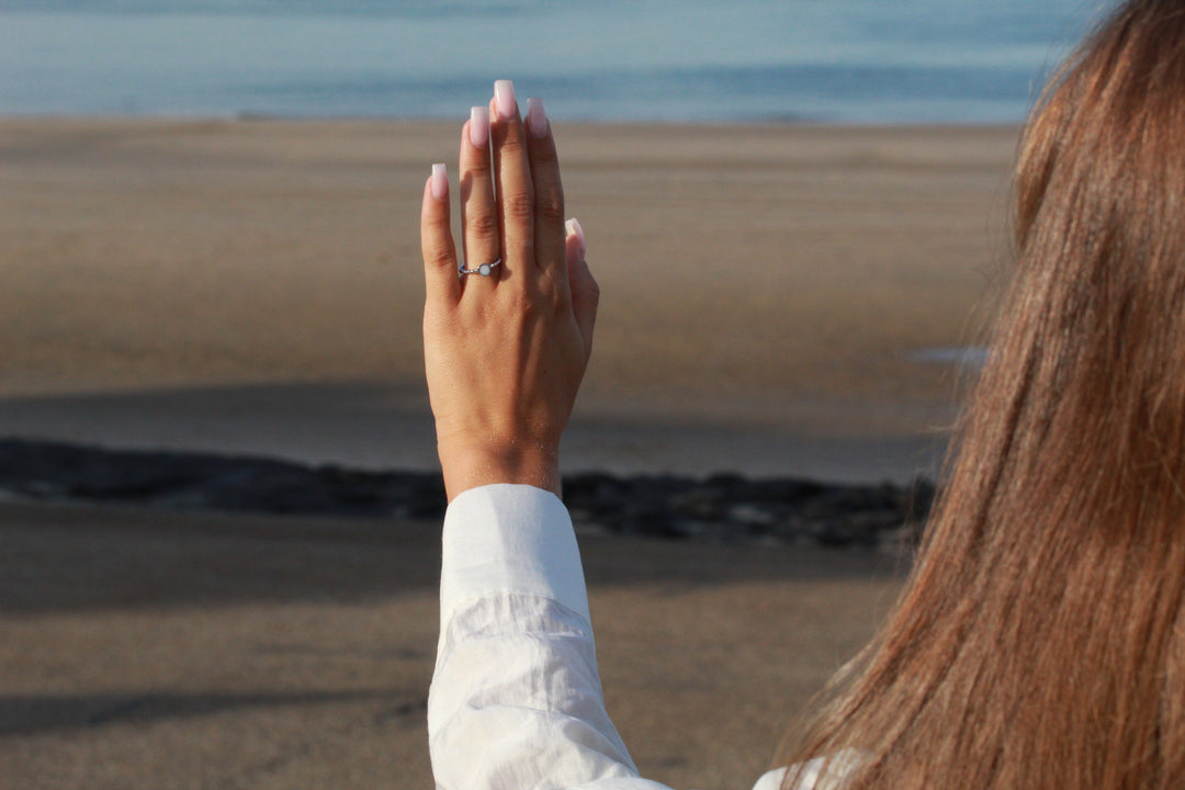 De hand van een vrouw versierd met de See You Isabelle | AsRing Zilver is opgeheven, tegen een achtergrond van een zandstrand en de oceaan. Ze draagt een wit shirt met lange mouwen en haar lange bruine haar is gedeeltelijk zichtbaar.